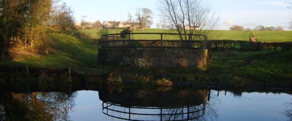 A circular stone bridge over a calm river, surrounded by grassy fields and trees.