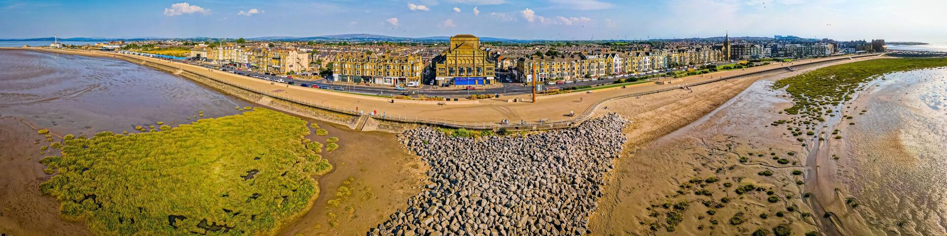 A coastal cityscape with a prominent historical building, a wide promenade, and a rocky shoreline.