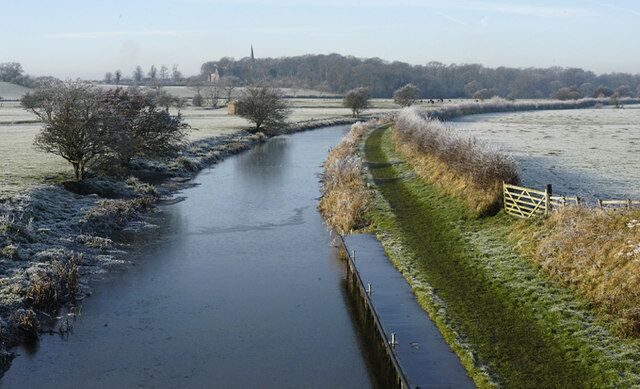 A frozen river with a narrow path alongside, surrounded by fields and trees.