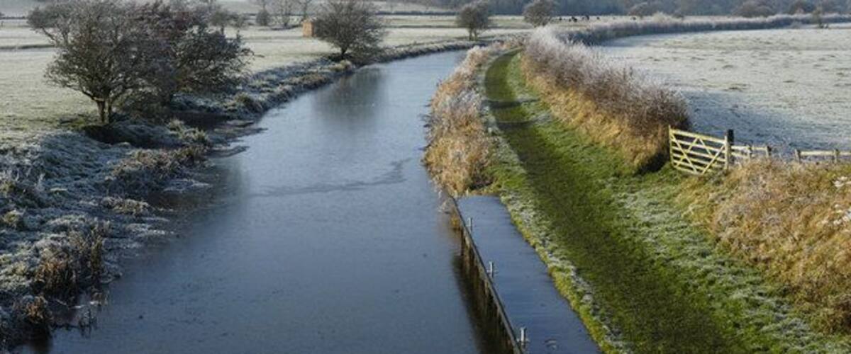A frozen river with a narrow path alongside, surrounded by fields and trees.