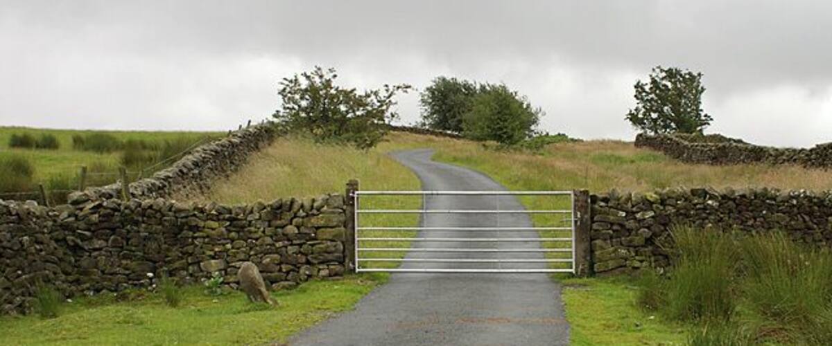 A rural road leading to a stone gate with a metal barrier.