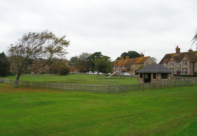 A grassy field with a fence, a small pavilion, and houses in the background.
