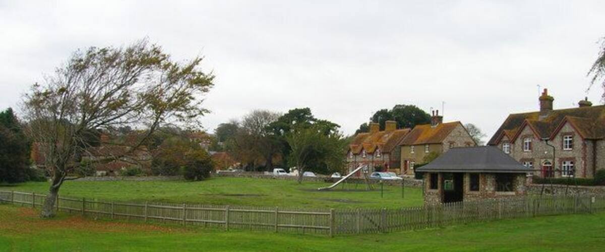 A grassy field with a fence, a small pavilion, and houses in the background.