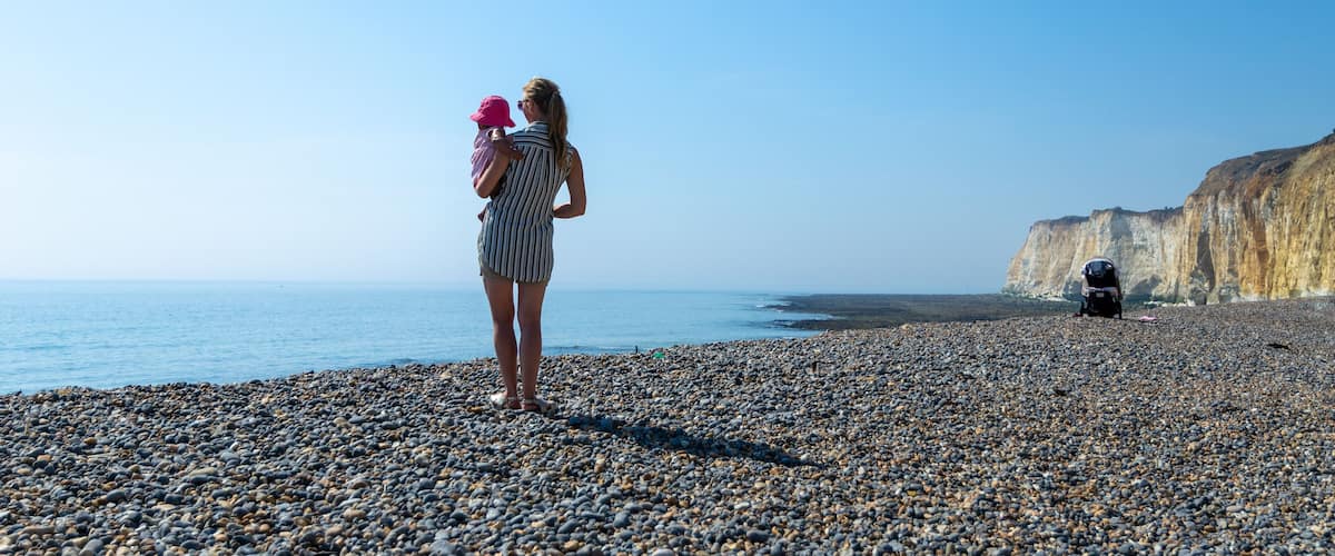 A pebbled beach with a person holding a child, a cliff in the background, and a clear sky.