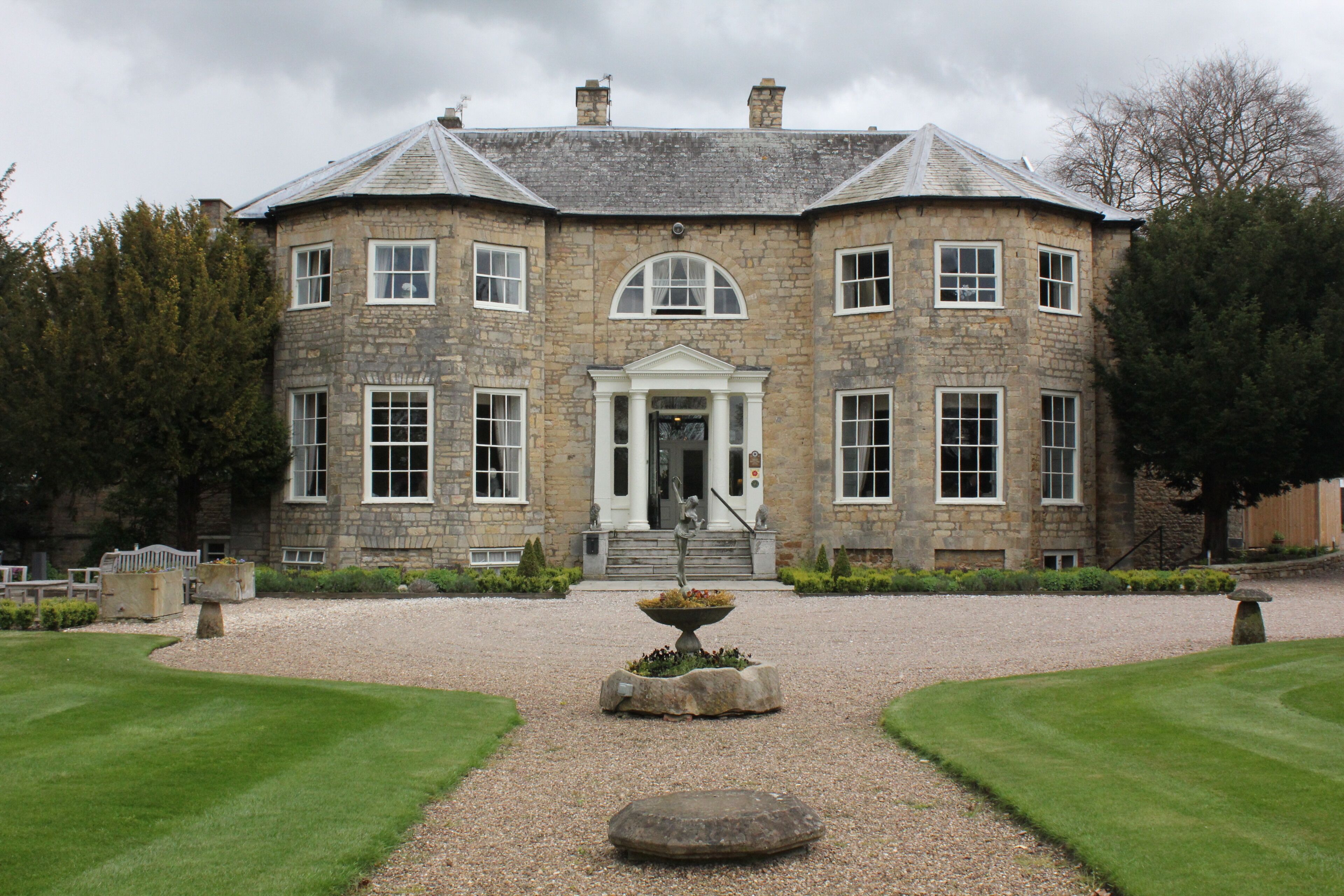 A large stone building with a symmetrical facade, a central entrance with a pediment, and a gravel driveway leading to a circular stone feature.