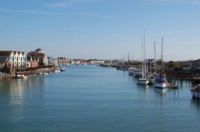 A calm harbor with boats docked along the pier, a large building with a dome on the left, and a clear blue sky.