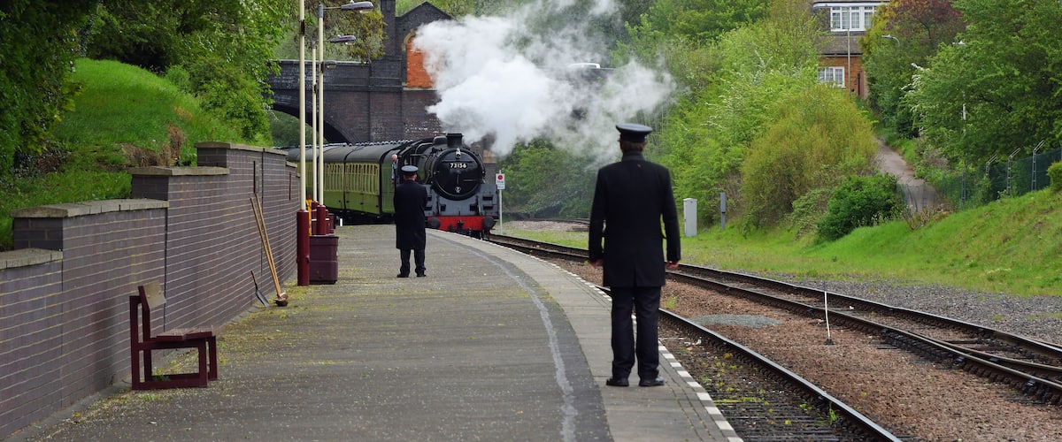 A steam train at a station with a person in uniform standing on the platform.