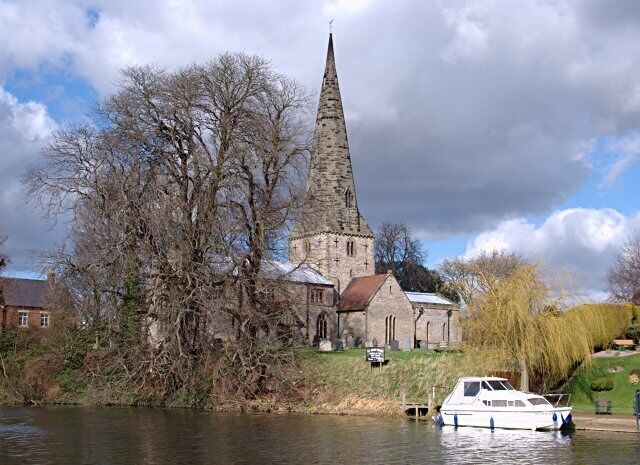 A church with a tall spire by a river, a boat docked near the shore, and leafless trees.