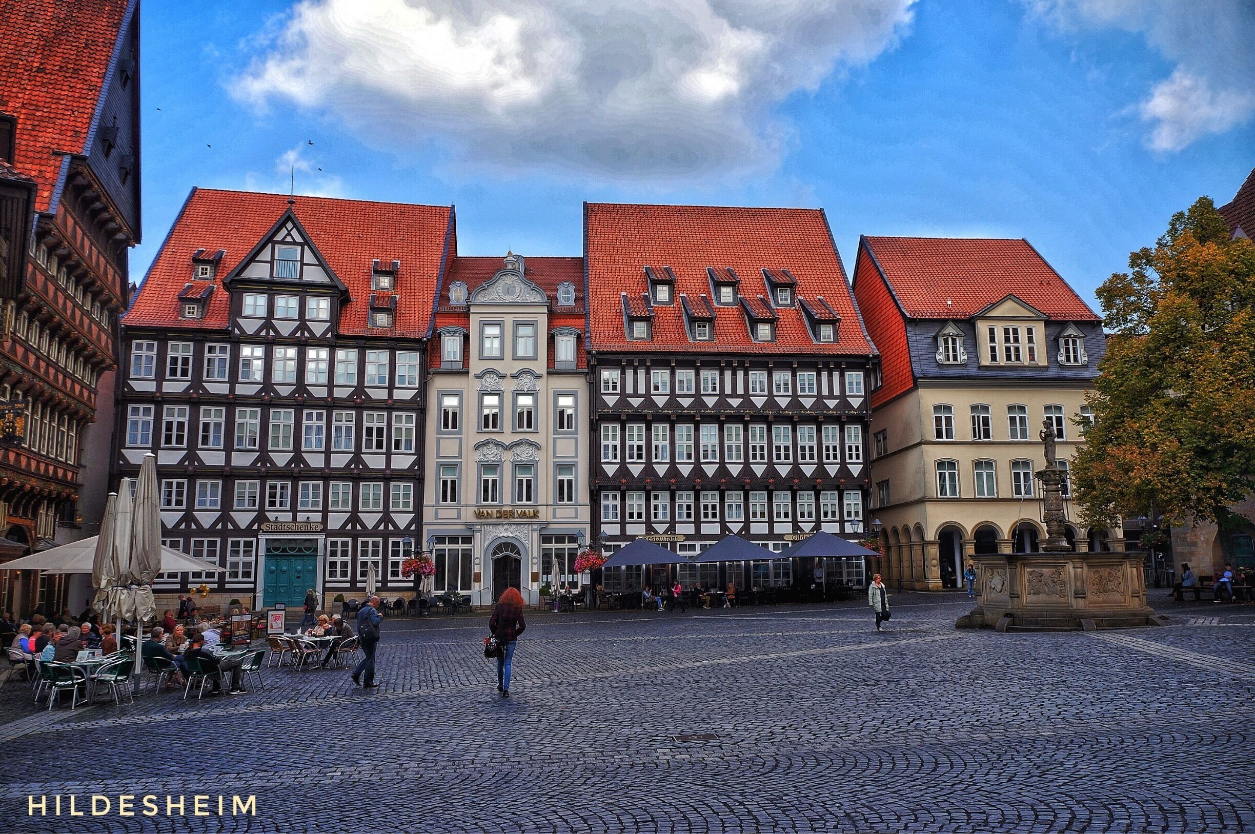 A cobblestone square with historic buildings, a fountain, and outdoor seating.