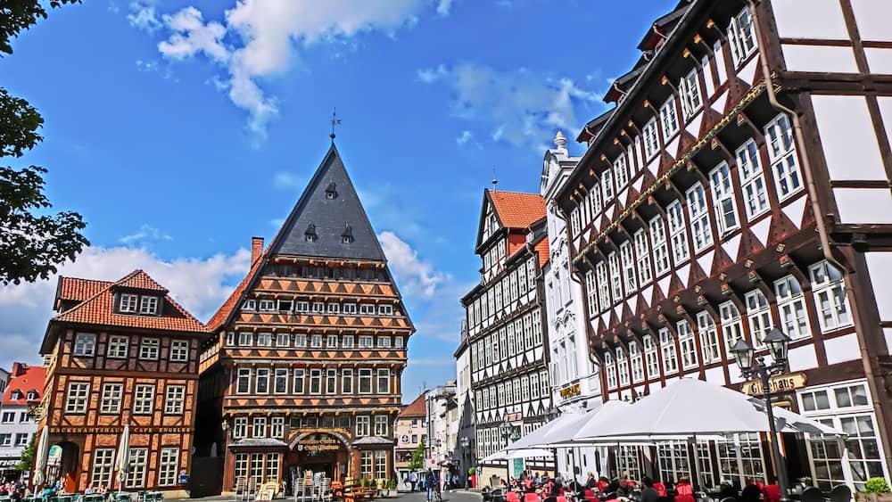 Ein Pflasterplatz mit traditionellen Fachwerkhäusern, einem markanten Kirchturm und Außenbereichen mit Sitzgelegenheiten.