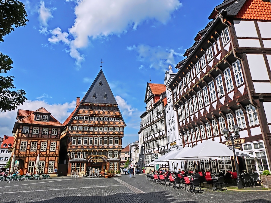 Ein Pflasterplatz mit traditionellen Fachwerkhäusern, einem markanten Kirchturm und Außenbereichen mit Sitzgelegenheiten.