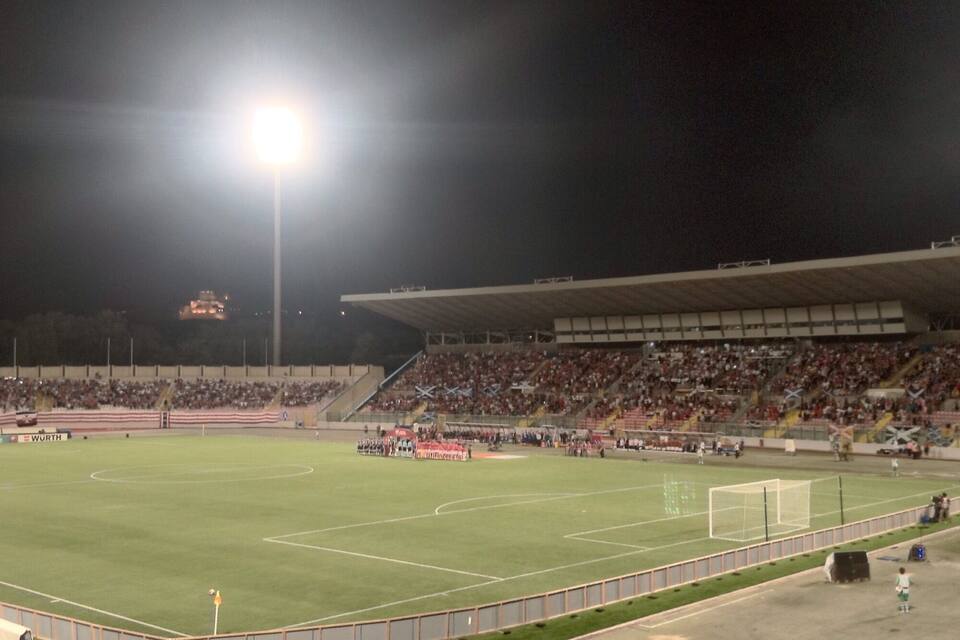 A football stadium at night with a large crowd, floodlights, and a goalpost.