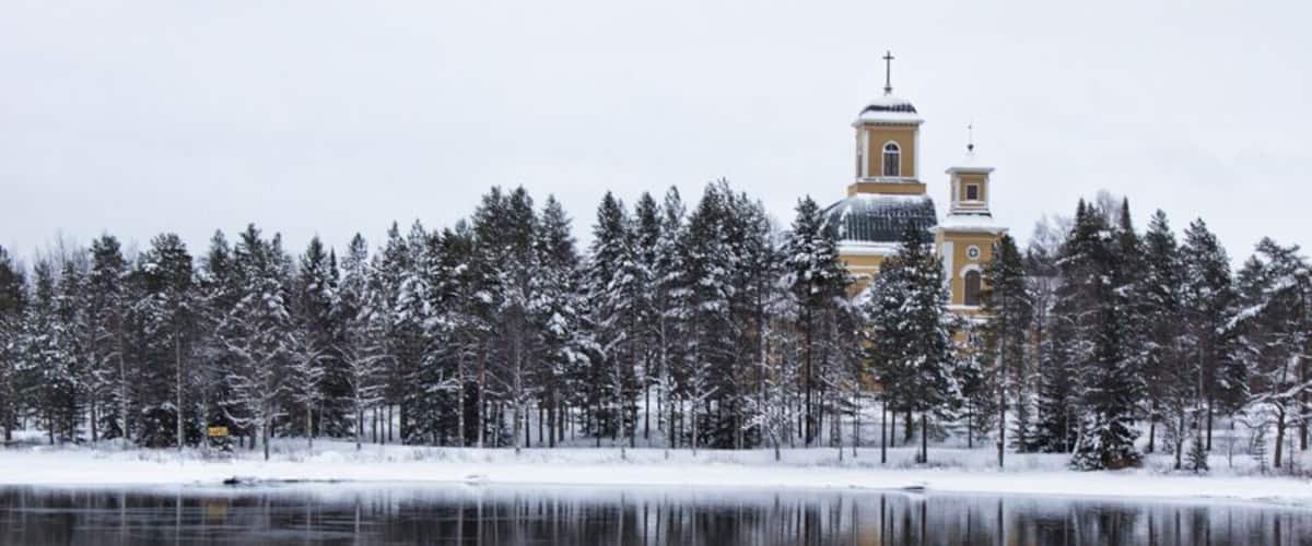 A snow-covered landscape with a church reflecting in a calm body of water.
