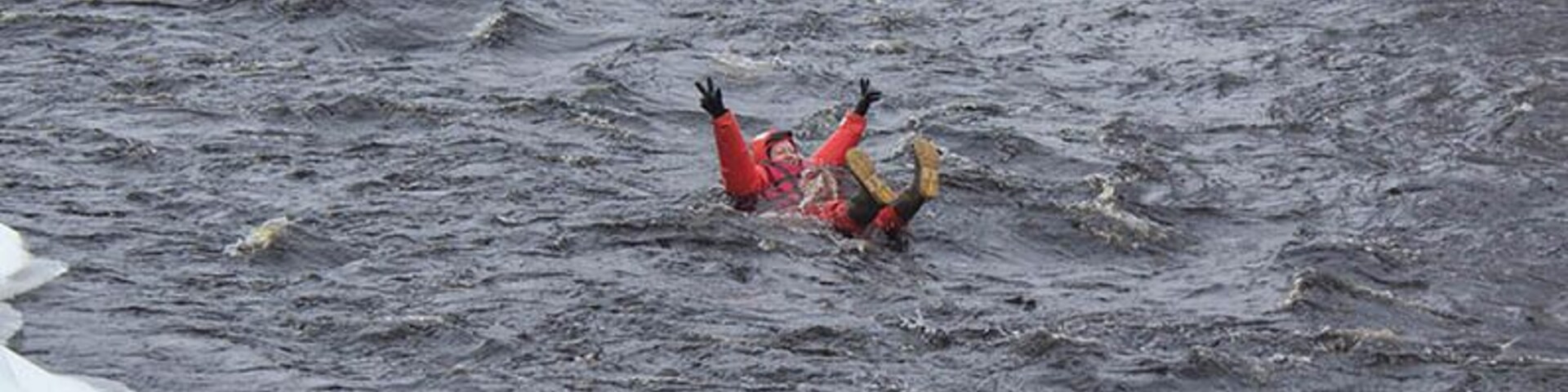 Una persona con una chaqueta roja y botas amarillas, parcialmente sumergida en un río con hielo.