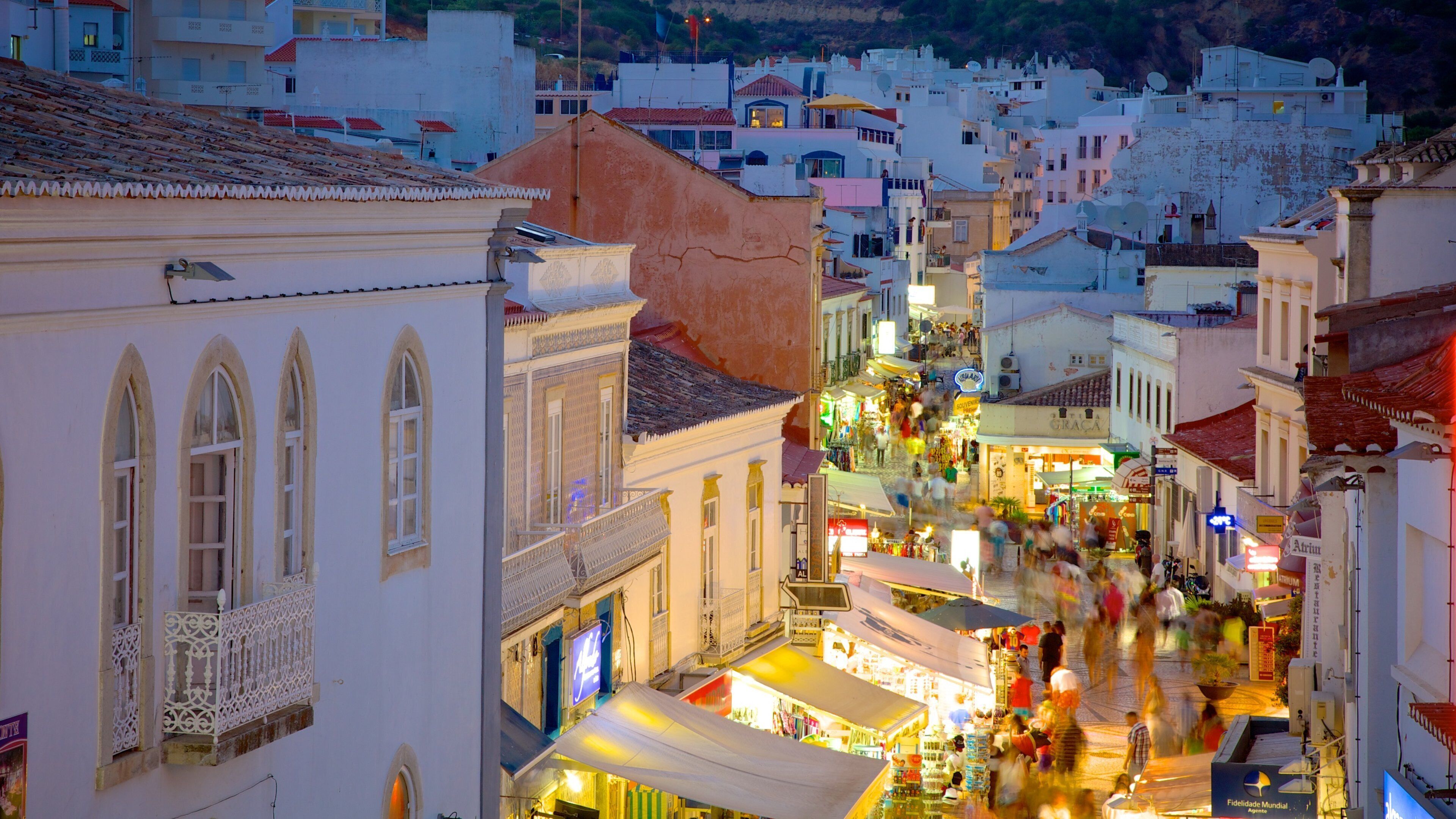 A bustling street in a historic town with white buildings, a market area, and a hillside in the background.