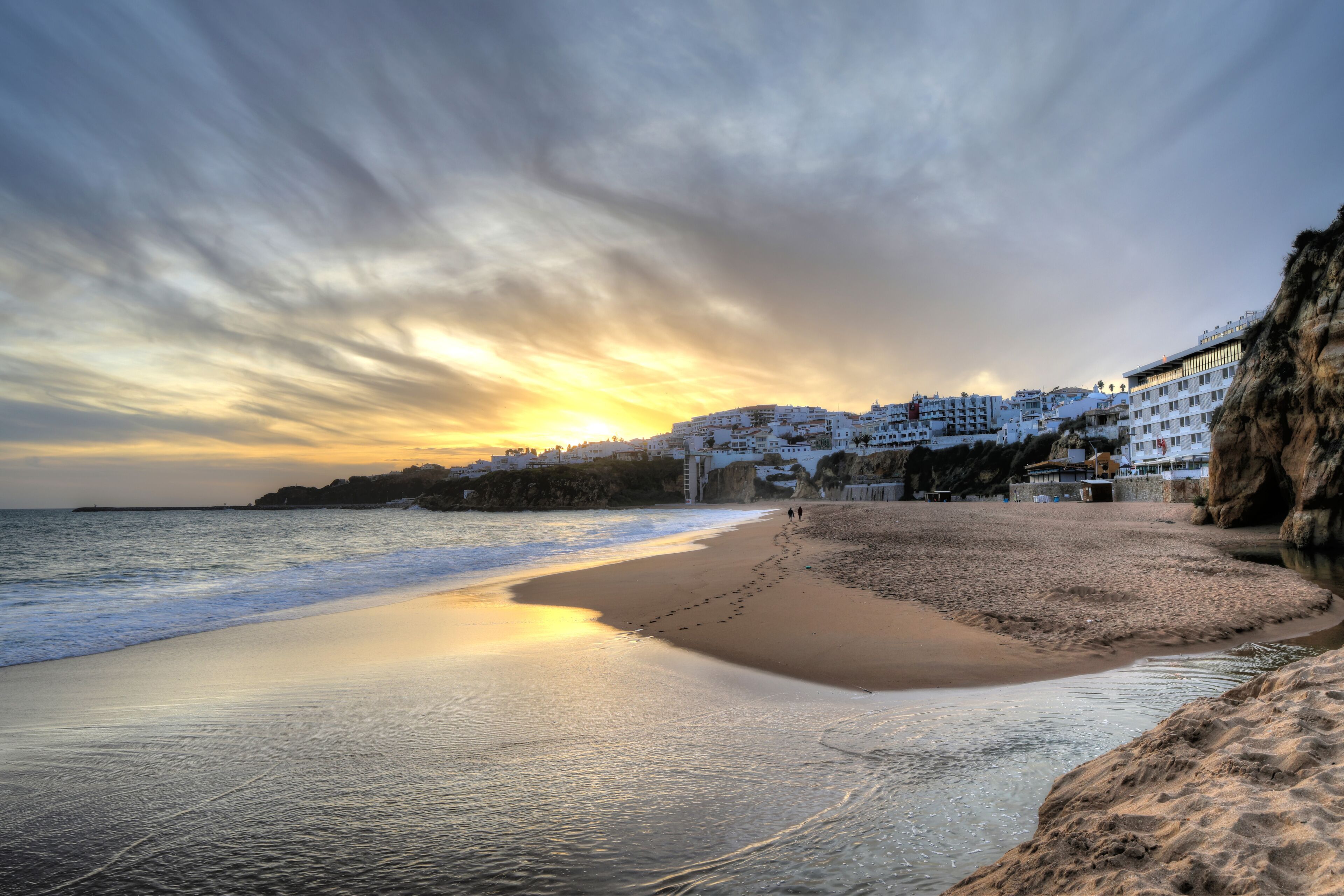 A beach with a sunset, calm waters, and white buildings on a cliff.
