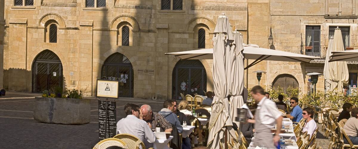 A stone building with arched windows and doors, outdoor seating with umbrellas, and a signboard with text.