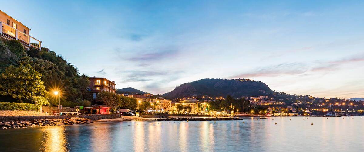A calm waterfront at dusk with illuminated buildings, a wooden dock, and a hill in the background.
