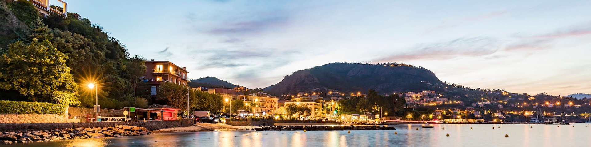 A calm waterfront at dusk with illuminated buildings, a wooden dock, and a hill in the background.
