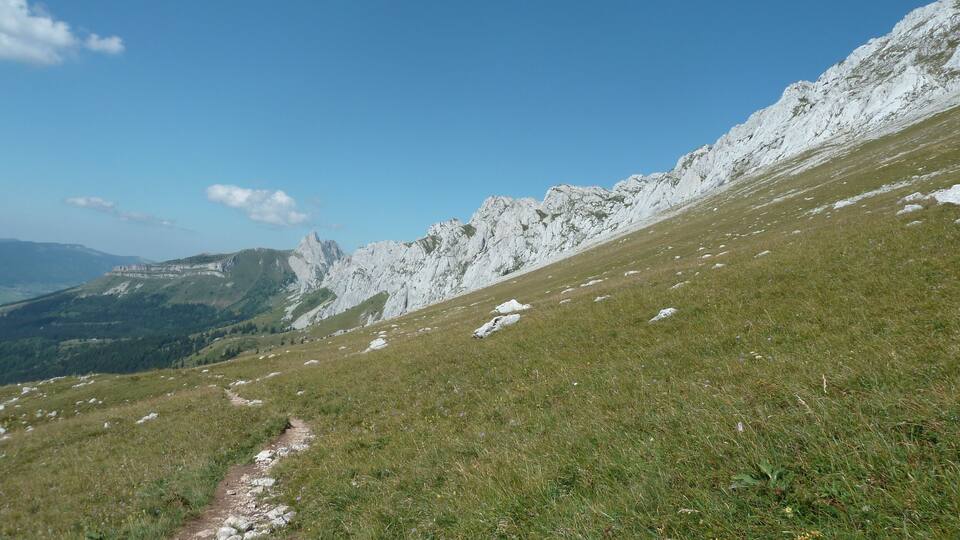 A grassy mountain path with rocky outcrops and distant mountains under a clear sky.