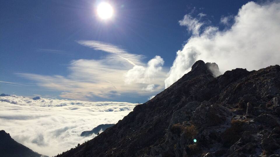 A mountain peak with a clear sky, sun, and clouds.
