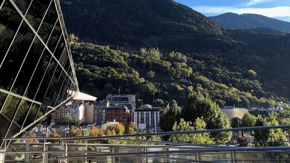 A modern bridge with glass panels, a mountainous landscape, and buildings in the background.