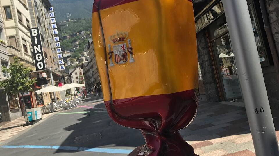 A sculpture of a red and yellow object with a coat of arms on a pedestal in an urban setting.