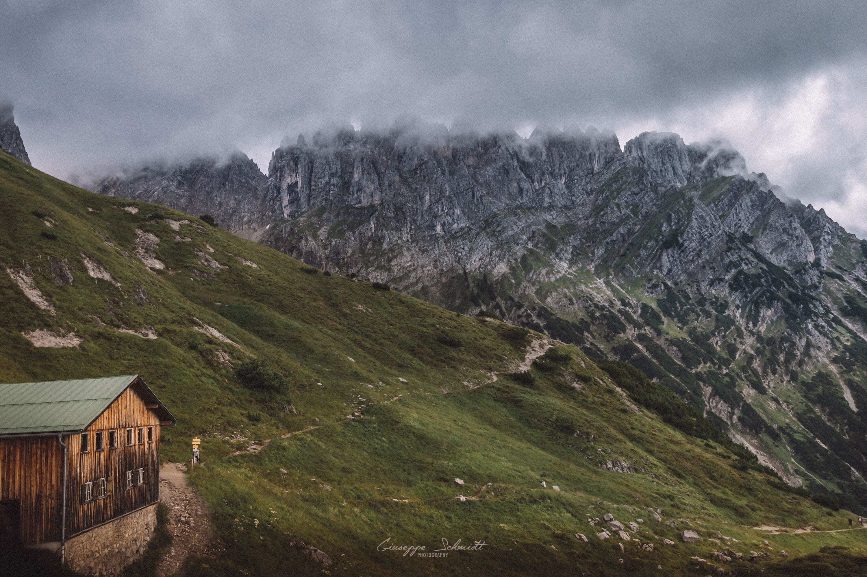 A wooden cabin on a grassy hillside with a mountain range in the background.