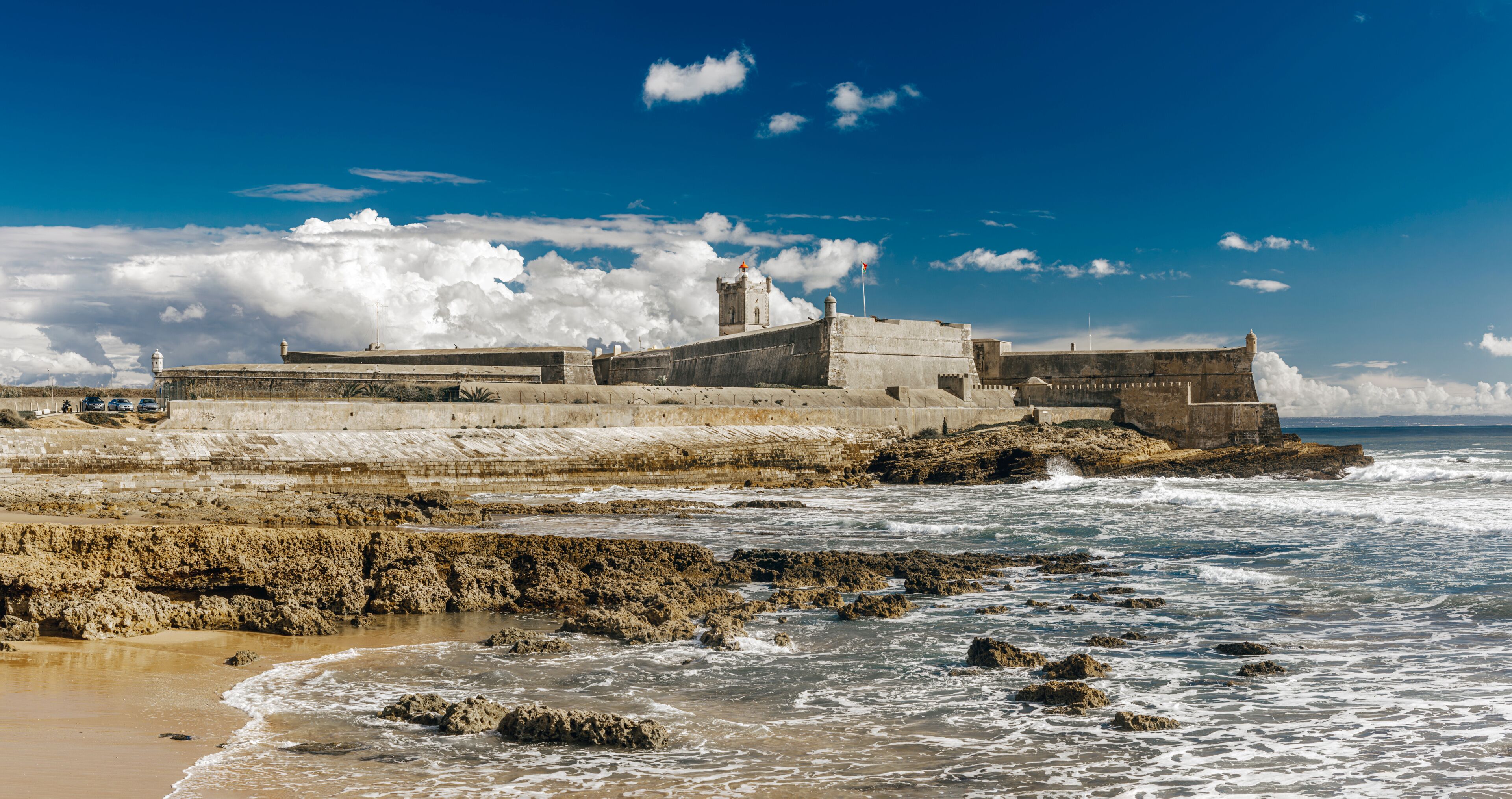 A coastal fortification with a prominent tower, waves crashing against the shore, and a clear blue sky.