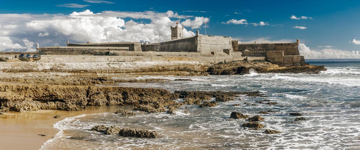 A coastal fortification with a prominent tower, waves crashing against the shore, and a clear blue sky.