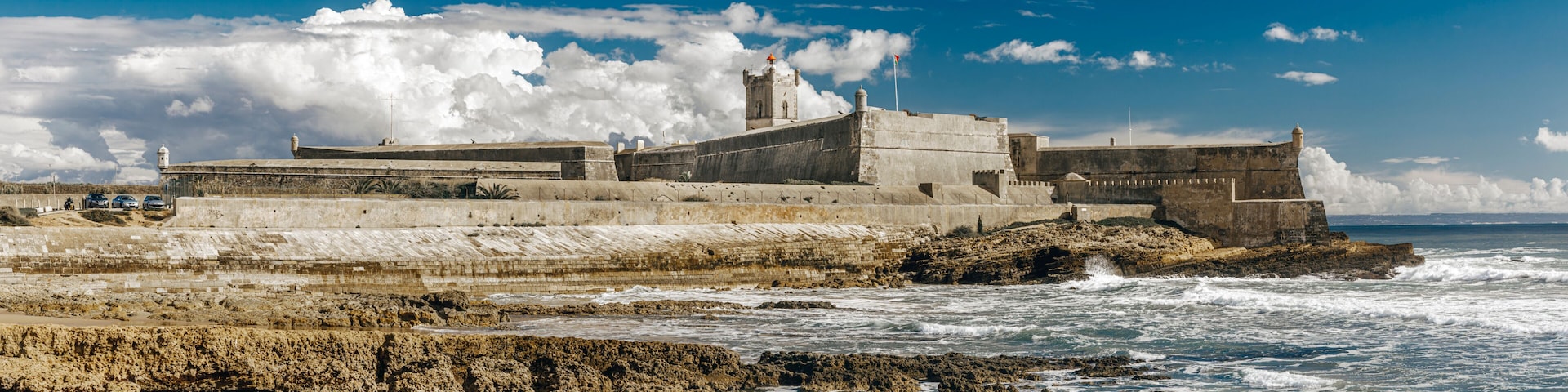 A coastal fortification with a prominent tower, waves crashing against the shore, and a clear blue sky.