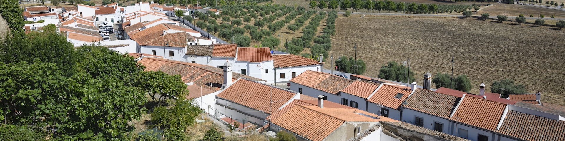 Un pueblo costero con edificios de tejados de teja, olivares y un paisaje montañoso.