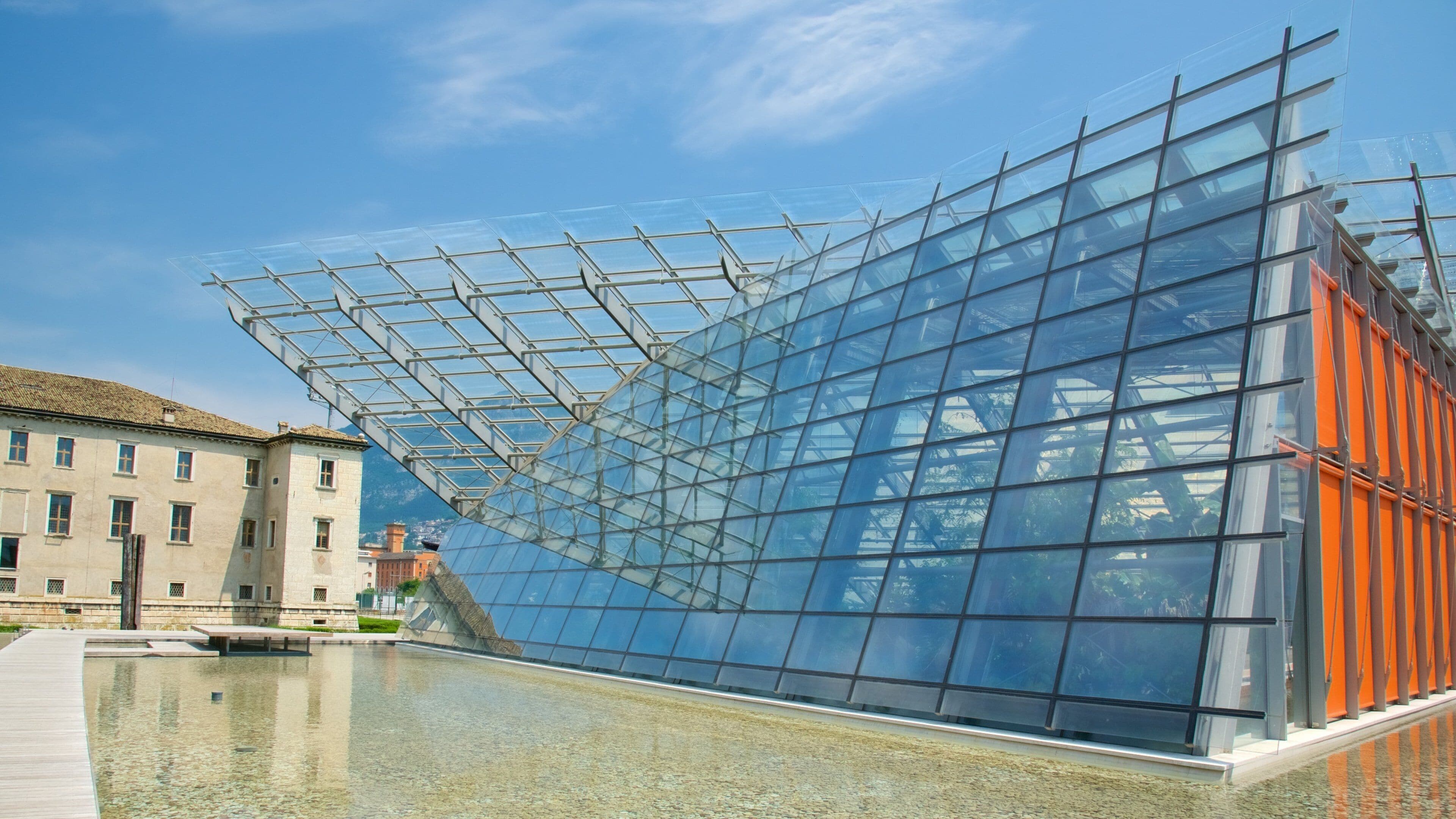 A modern glass structure with a metal framework, a reflective water surface, and a historic building in the background.