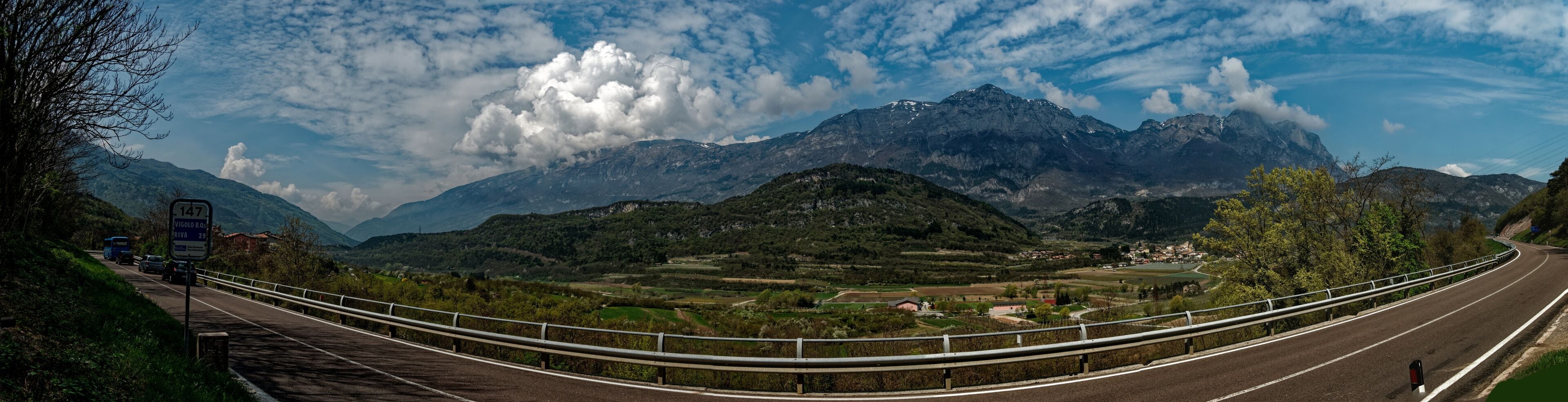 A winding road with a guardrail, surrounded by lush green hills and mountains under a partly cloudy sky.