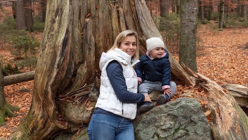 A woman and a child sitting on a large rock in a forest with a prominent tree trunk in the background.