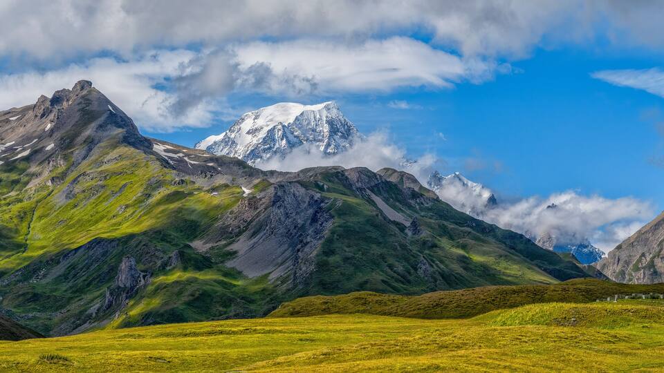 A mountain range with green valleys and a snow-capped peak under a partly cloudy sky.