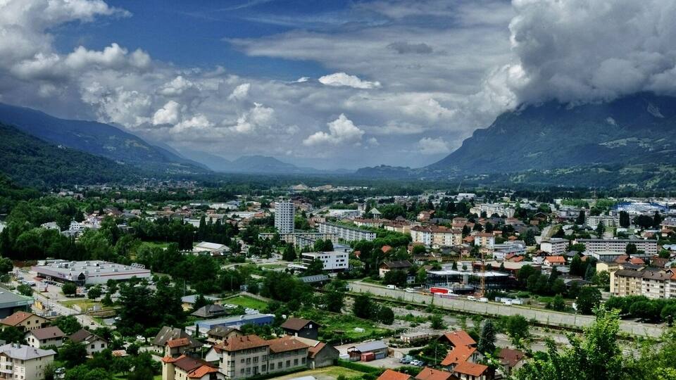 A town nestled in a valley with mountains in the background, surrounded by greenery and featuring a mix of residential and commercial buildings.