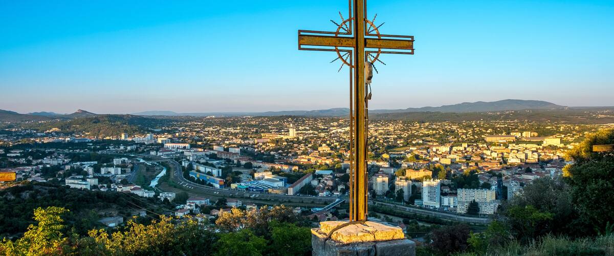 A cross on a hill overlooking a cityscape with a river and mountains in the background.