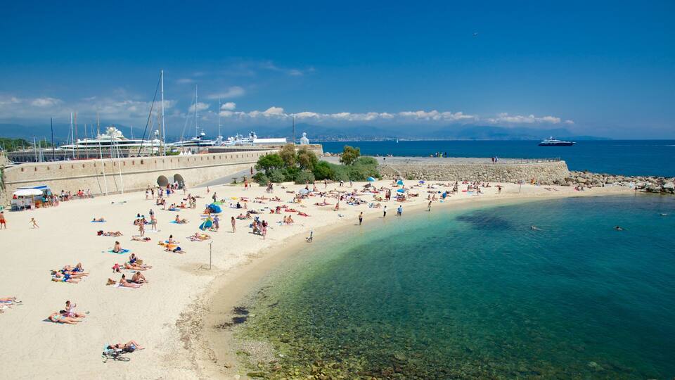 A crowded beach with clear water, boats, and a stone wall.