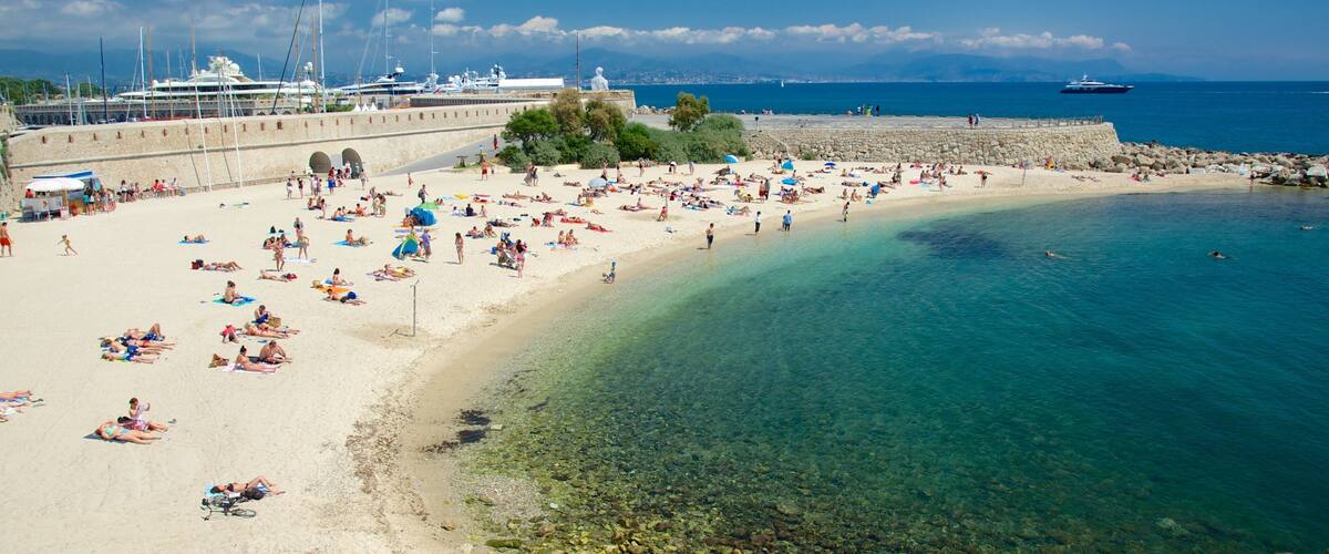 A crowded beach with clear water, boats, and a stone wall.
