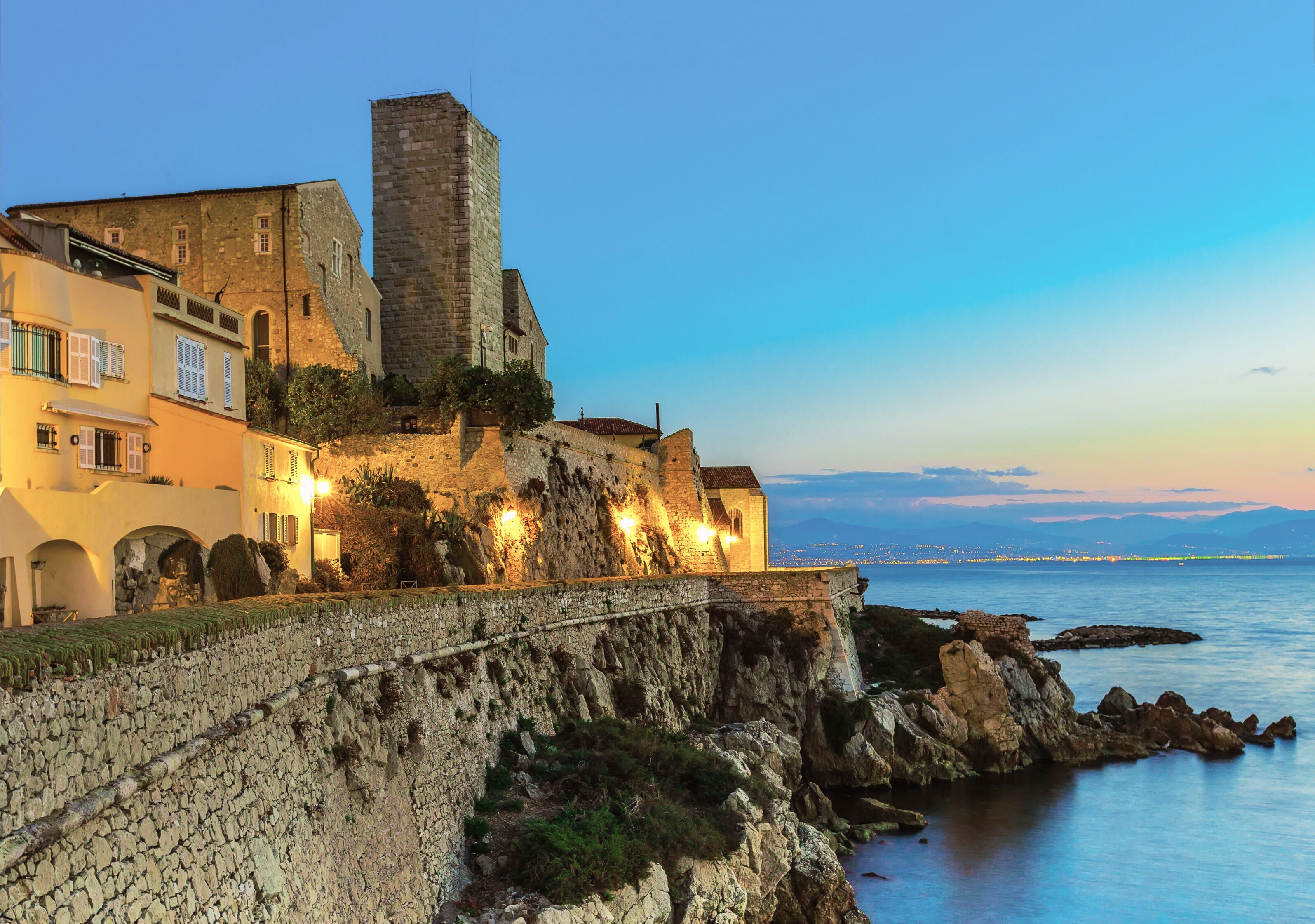 A coastal town with historic stone buildings, a prominent tower, and a stone wall overlooking the sea.