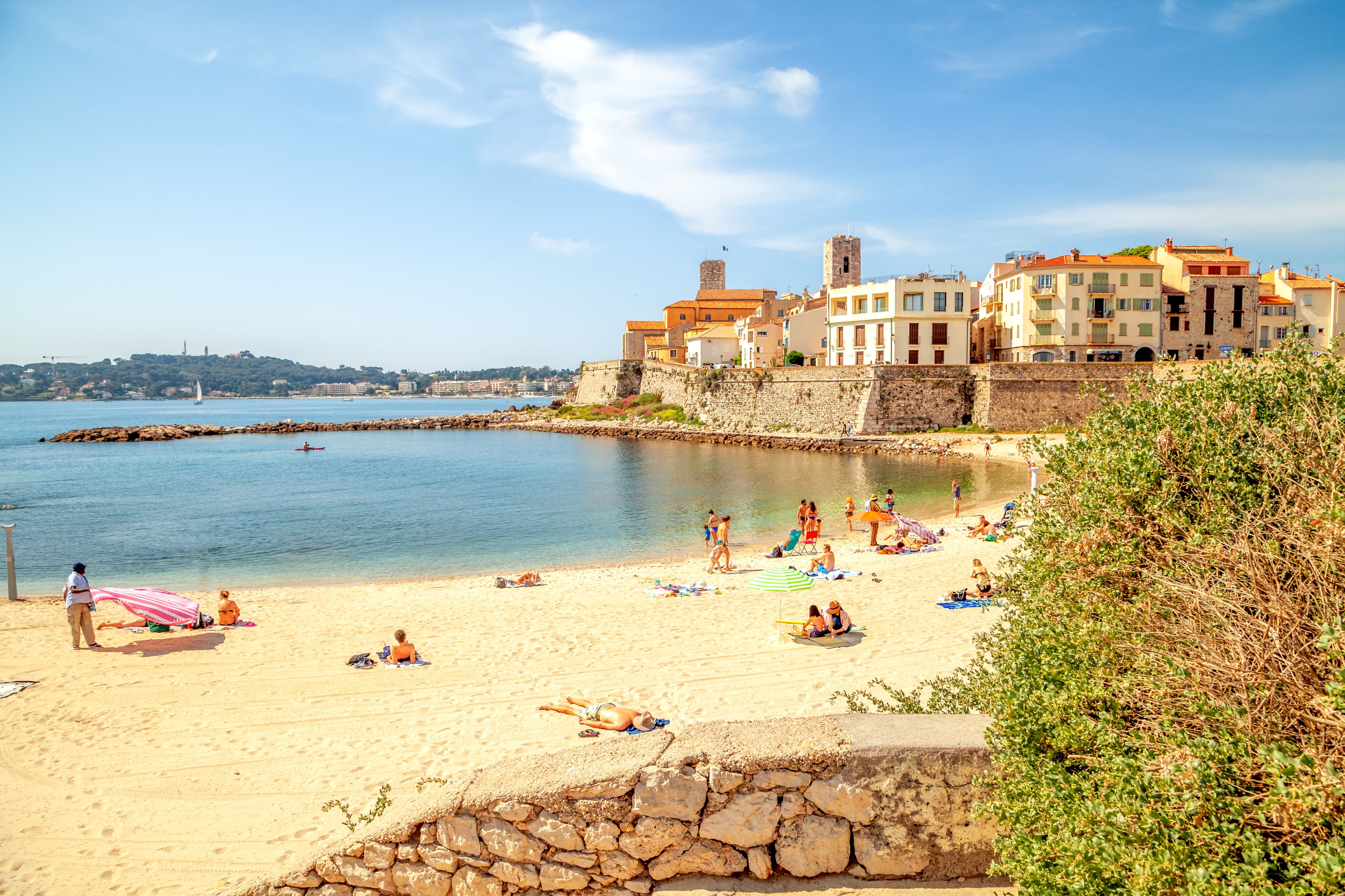 A sunny beach with people sunbathing, a clear blue sea, and historic buildings in the background.