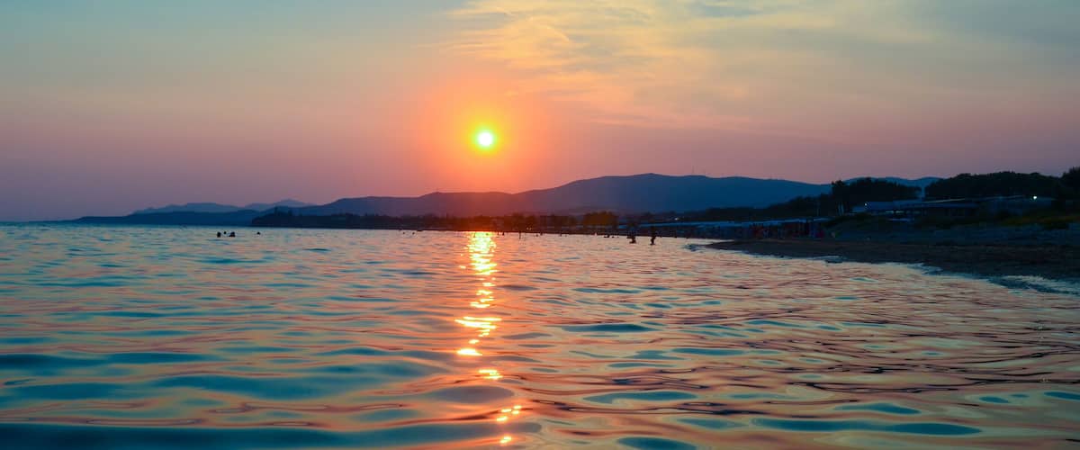 A sunset over a calm sea with reflections, a distant shoreline, and mountains in the background.