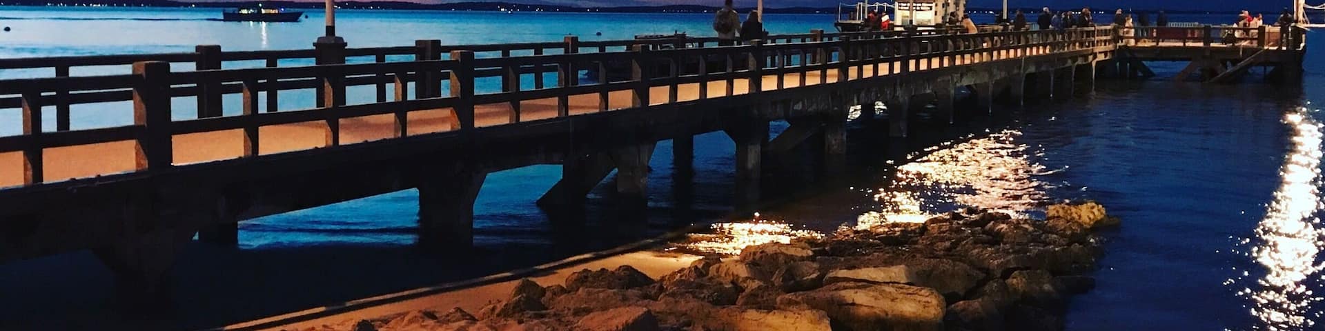 A beach at dusk with a wooden pier extending into the water, illuminated by street lamps.
