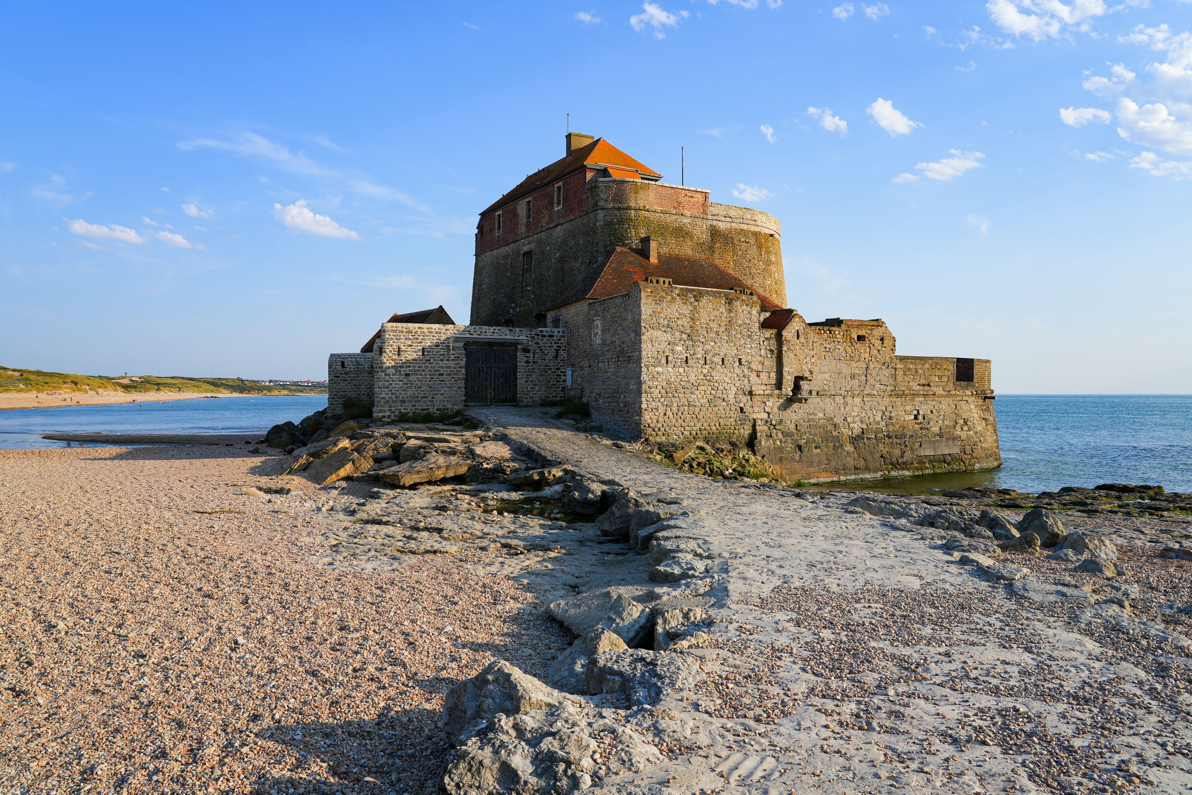 Una fortaleza de piedra histórica junto a una playa de guijarros, con el mar al fondo.