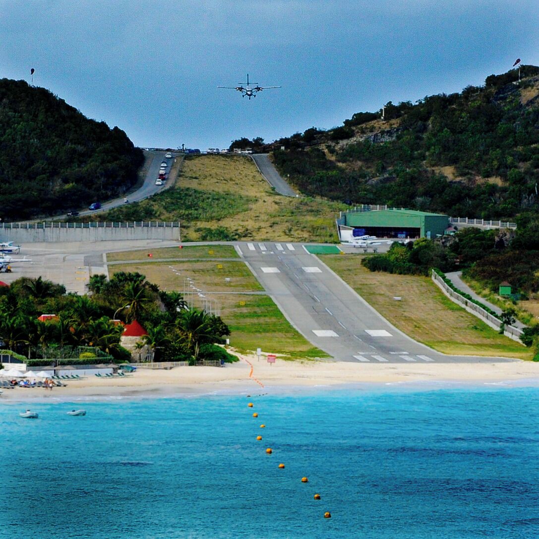 Uma estrada costeira que leva a uma praia, com céu claro e um avião ao longe.
