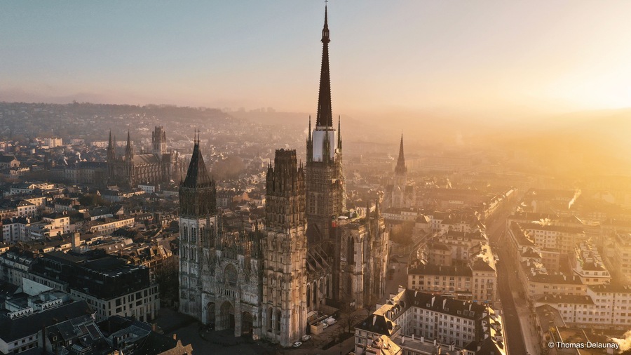 Une cathédrale dotée d’une haute flèche, entourée d’un paysage urbain composé de différents bâtiments et surplombée d’un ciel dégagé.