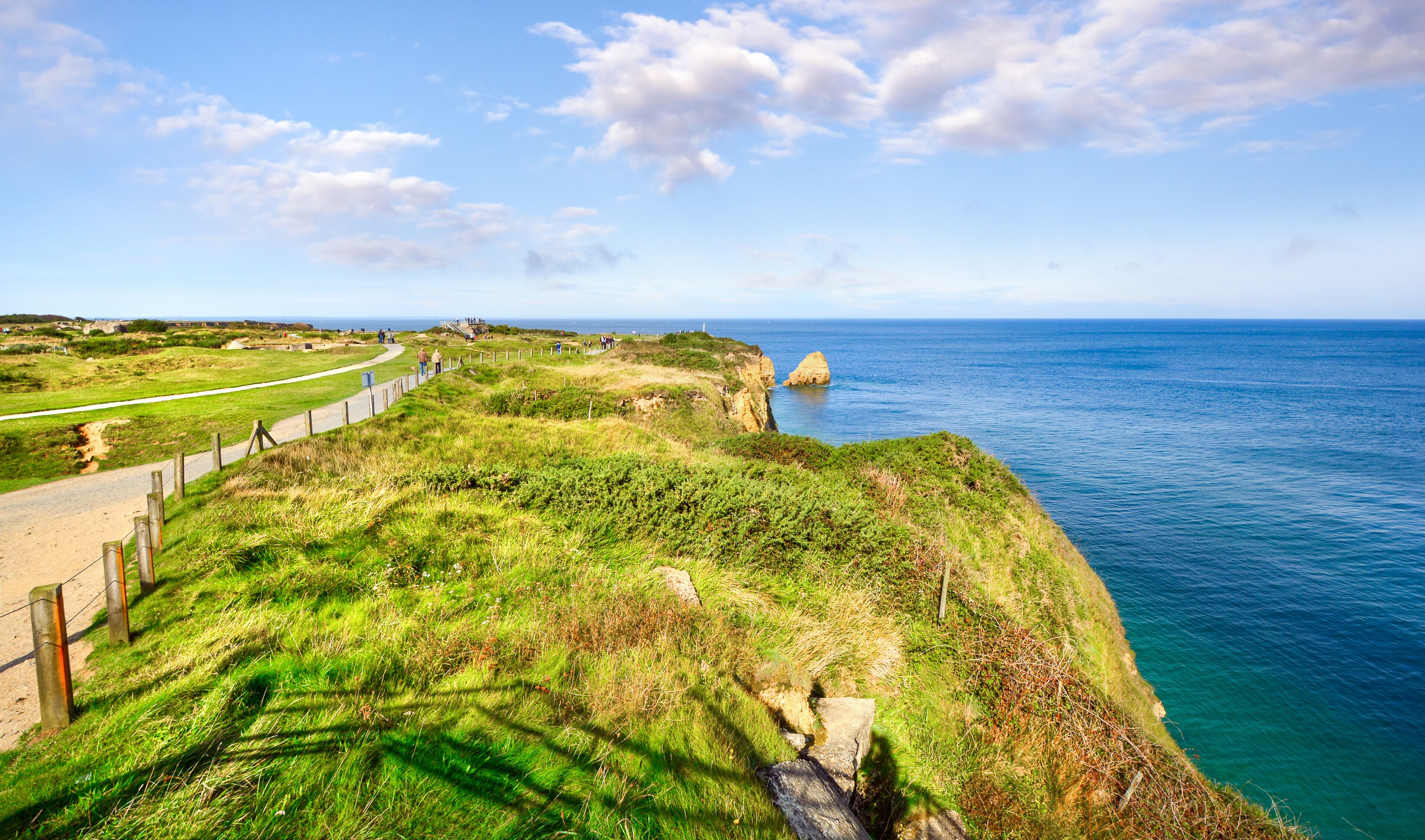 A coastal path with a grassy cliff edge, a clear blue sea, and a distant lighthouse.