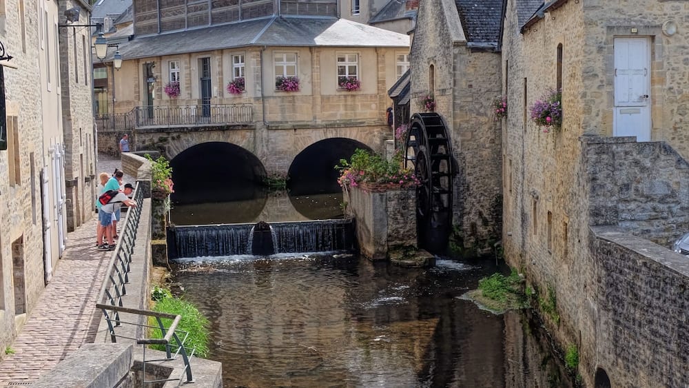 Ein schmaler Kanal mit Wassermühle und Brücke, umgeben von historischen Steinbauten und einem Kirchturm im Hintergrund.