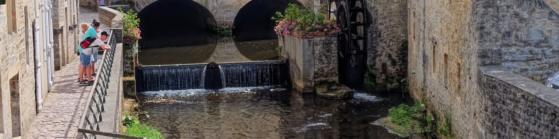 A narrow canal with a watermill and a bridge, surrounded by historic stone buildings and a church spire in the background.